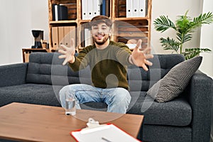 Arab man with beard working on depression at therapy office looking at the camera smiling with open arms for hug