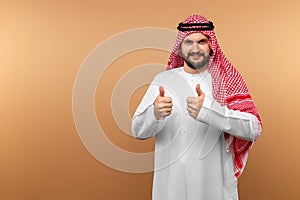 Arab man businessman in national clothes shows thumbs up, like, beige background. Dishdasha, kandora, thobe, middle east photo
