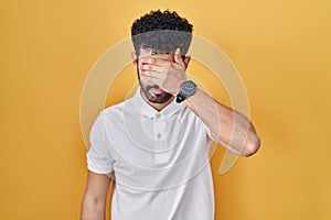 Arab man standing over yellow background covering eyes with hand, looking serious and sad