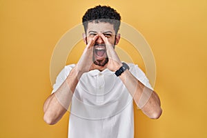 Arab man standing over yellow background shouting angry out loud with hands over mouth