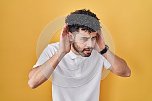 Arab man standing over yellow background trying to hear both hands on ear gesture, curious for gossip