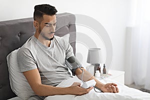 Arab Man Using Upper Arm Blood Pressure Monitor While Sitting In Bed