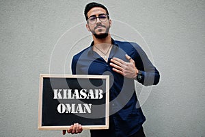 Arab man wear blue shirt and eyeglasses hold board with Khasab Oman inscription. Largest cities in islamic world concept