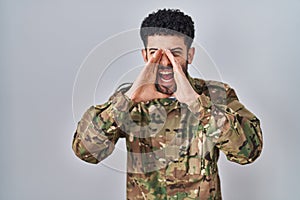 Arab man wearing camouflage army uniform shouting angry out loud with hands over mouth