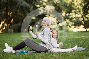 Arab mom and her daughter sitting on gray carrymat in the summer park, holding bottles with water