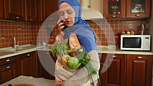 Arab Muslim woman in hijab, talks on mobile phone while putting a shopping bag with organic food on table in kitchen