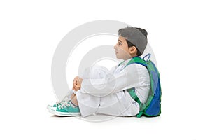 Arab school boy sitting on ground with a smile on his face, wearing white traditional Saudi Thobe, back pack and sneakers, raising photo