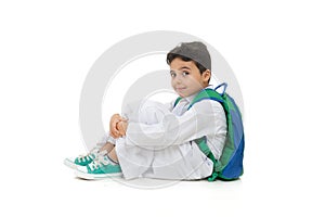 Arab school boy sitting on ground with a smile on his face, wearing white traditional Saudi Thobe, back pack and sneakers, raising photo
