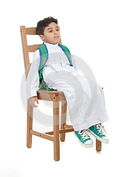 Arab school boy sitting sleepy and tired on a wooden chair, wearing white traditional Saudi Thobe, back pack and sneakers, raising photo