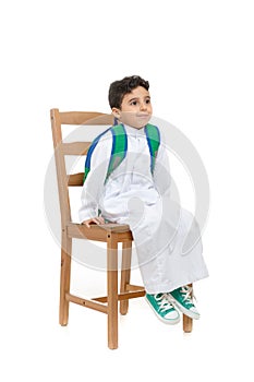 Arab school boy sitting on a wooden chair with a smile on his face, wearing white traditional Saudi Thobe, back pack and sneakers photo