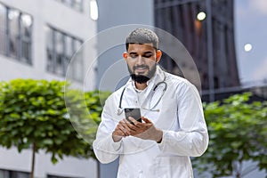 Arab smiling young male doctor, student standing near university hospital in uniform and using phone