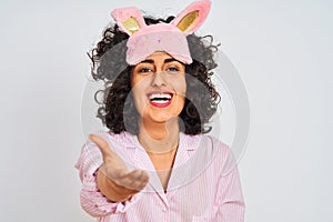 Arab woman with curly hair wearing pajama and sleep mask over isolated white background smiling friendly offering handshake as