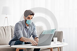 Arab young business man working from home with laptop, wearing protective medical face mask, sitting on sofa, free space