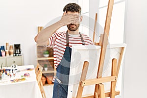Arab young man at art studio covering eyes with hand, looking serious and sad