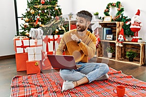 Arab young man using laptop sitting by christmas tree pointing to the back behind with hand and thumbs up, smiling confident