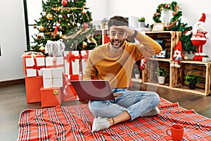 Arab young man using laptop sitting by christmas tree very happy and smiling looking far away with hand over head