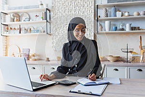 Arab young woman in hijab and burqa works online as accountant, auditor from home. Remotely works on a laptop, with
