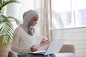 Arab young woman in hijab sitting on sofa at home, holding laptop on lap, looking worriedly at screen and spreading