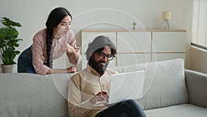 Arabian Indian couple on couch use laptop computer shopping online together pay bills order food at home choose buying