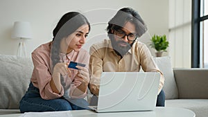 Arabian Indian muslim couple family sit on sofa in living room home office use credit bank card typing code number in