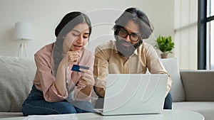 Arabian Indian muslim couple family sit on sofa in living room home office use credit bank card typing code number in