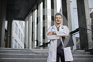 Arabian male physician, holding clipboard, standing with arms crossed outdoors clinic