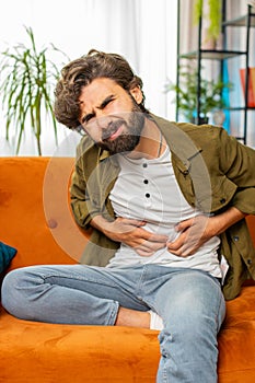 Arabian man sits on sofa feeling sudden strong abdominal stomach ache, gastritis problem, diarrhea