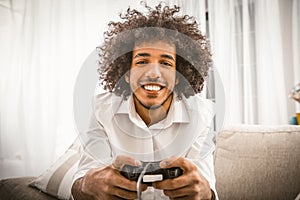 Arabic gay playing computer game. Young man holds Play Station joystick sitting at sofa in backlit. Smiling handsome