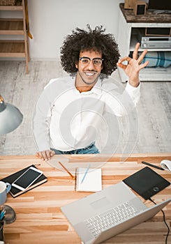 Arabic handsome shows OK gesture smiling at camera. Happy man in glasses working at home workpace with laptop, mobile