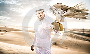 Arabic man with traditional emirates clothes walking in the desert with his falcon bird