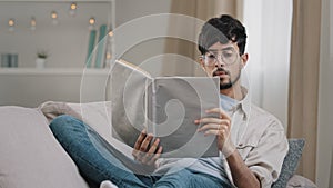 Arabic young smart guy student bearded man in glasses lies on sofa at home reading book textbook learning new language