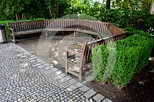 Arch of the flowerbed surround the wooden bench in the park. The bench is bent like a horseshoe, with a plank surface by the sidew