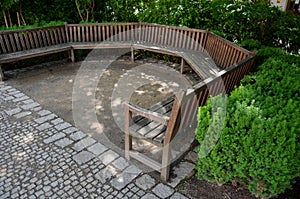 Arch of the flowerbed surround the wooden bench in the park. The bench is bent like a horseshoe, with a plank surface by the sidew