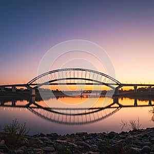 Arched bridge gracefully spans a tranquil river at sunset. The bridge's reflection