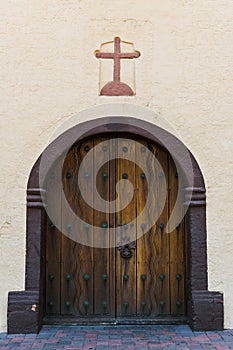 Arched rustic wood double doors under a cross in a plastered adobe wall of an old Spanish mission church