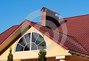 Arched window and brick chimney on the red metal tile roof under blue sky, house exterior