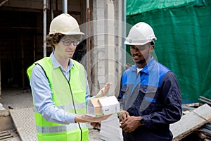 Architect and builder talking and holding model house at construction site.