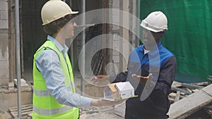 Architect and builder talking and holding model house at construction site.