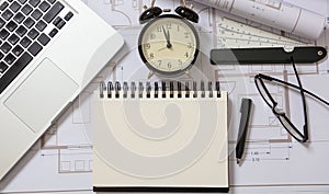 Architect engineer blank notebook, laptop and alarm clock on construction project blueprints background