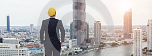 Architect man wear black suit and yellow helmet view from back standing on building looking for cityscape. looking for future