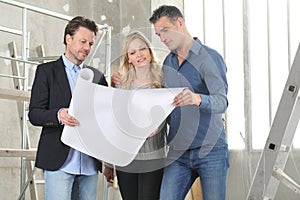 Architect showing house design plans to a young couple. House building goal concept. Meeting at interior construction site to talk