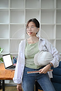 Architect woman holding with hard hat safety helmet and standing in office.