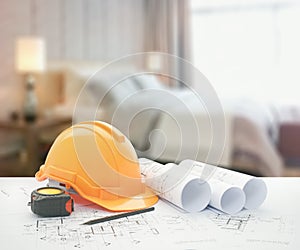 Architectural blueprint with safety helmet and tools over modern bedroom with decorative lamp as background