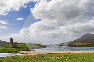 Ardvreck Castle ruins stand by tranquil water, hills of Scotland in distance under vast cloudy sky