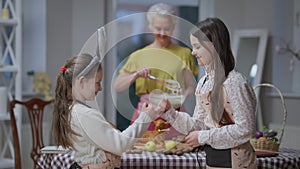 Argued sisters making peace gesturing pinky swear hugging indoors at home. Blurred smiling grandmother cooking Easter