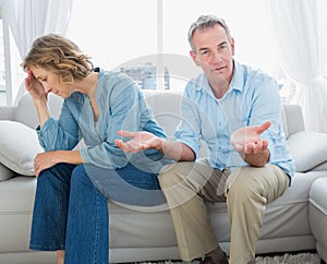 Arguing middle aged couple sitting on the couch with man gesturing at camera