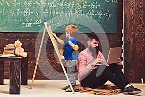 Arithmetic lesson at school. Kid writing on chalkboard while concentrated teacher works on laptop. Side view sitting man