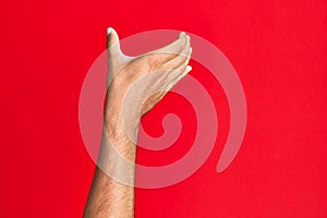 Arm of caucasian white young man over red isolated background holding invisible object, empty hand doing clipping and grabbing