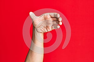 Arm of caucasian white young man over red isolated background holding invisible object, empty hand doing clipping and grabbing