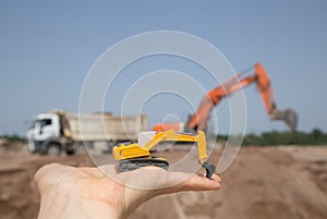 On the arm is a small orange toy excavator. Behind him, out of focus, is a real working excavator and dump truck